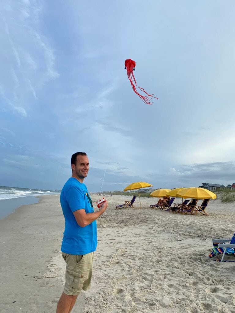 A man in a blue shirt smiles while flying a red kite shaped like a jellyfish on the sandy shore of Kure Beach, NC, with yellow beach umbrellas and the ocean in the background.