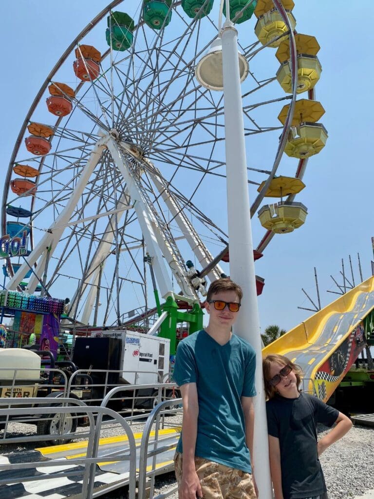 Two kids smiling in front of colorful Gondolas at the Carolina Beach Boardwalk, enjoying a sunny day of fun and rides.