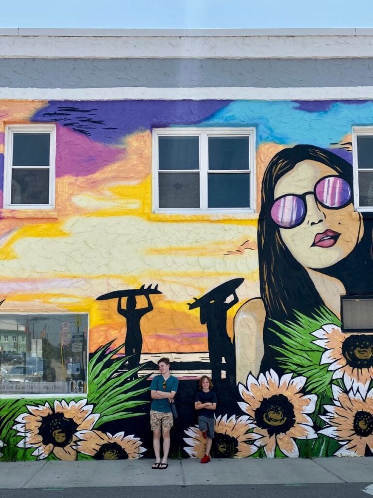A vibrant mural depicting a woman with sunglasses, surfers, and sunflowers, with two kids standing in front, located on the Carolina Beach Boardwalk.