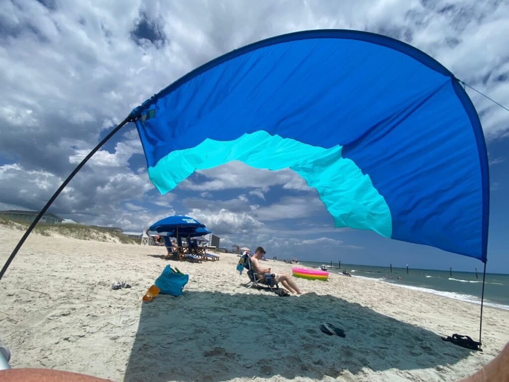 A vibrant blue Shibumi shade provides shade on the sandy shore of Kure Beach, NC, with ocean waves and beachgoers in the distance.