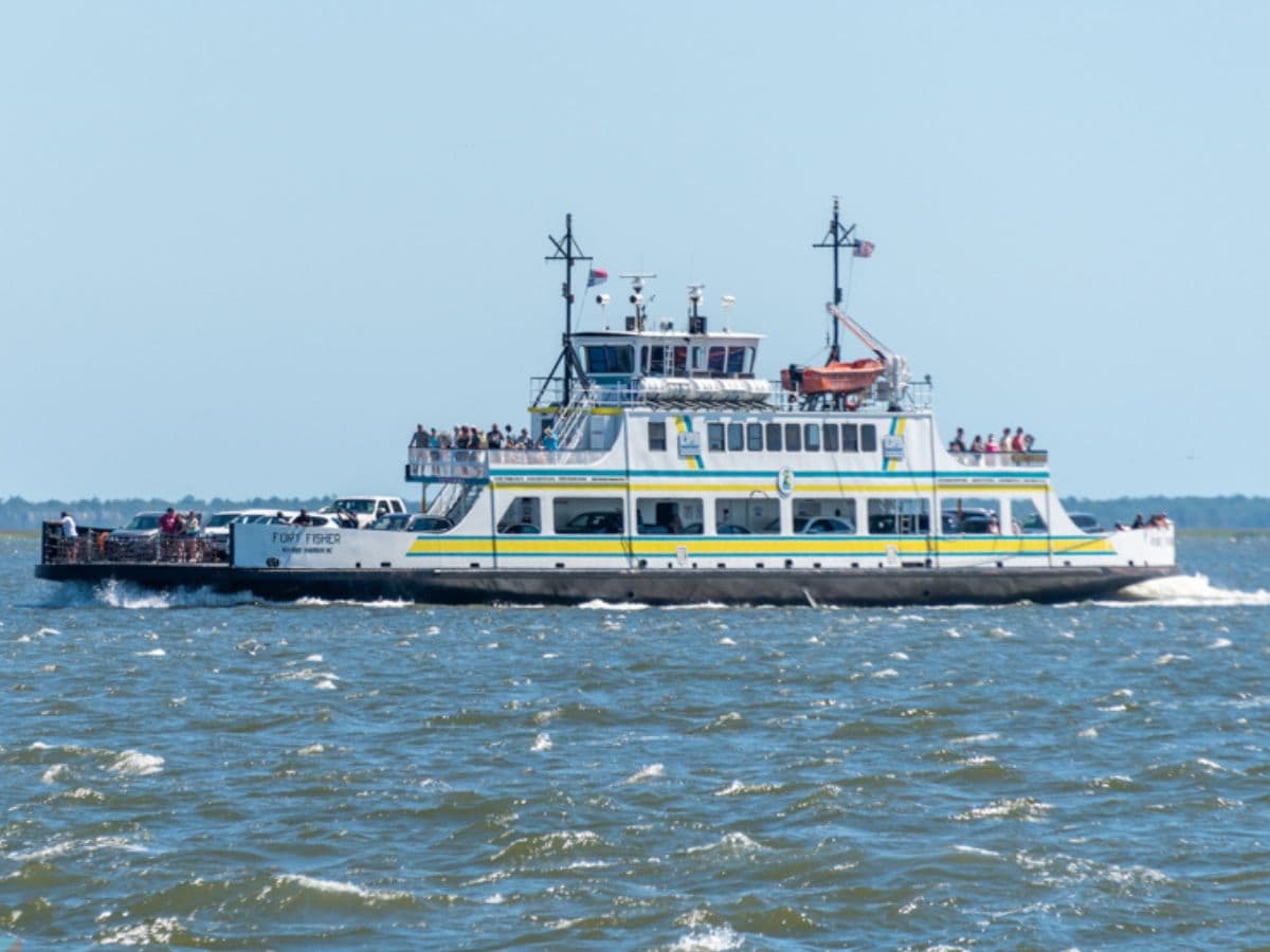 Fort Fisher Ferry carrying vehicles and passengers across the water on a sunny day near Kure Beach, NC, with a clear blue sky and gentle waves in the background.