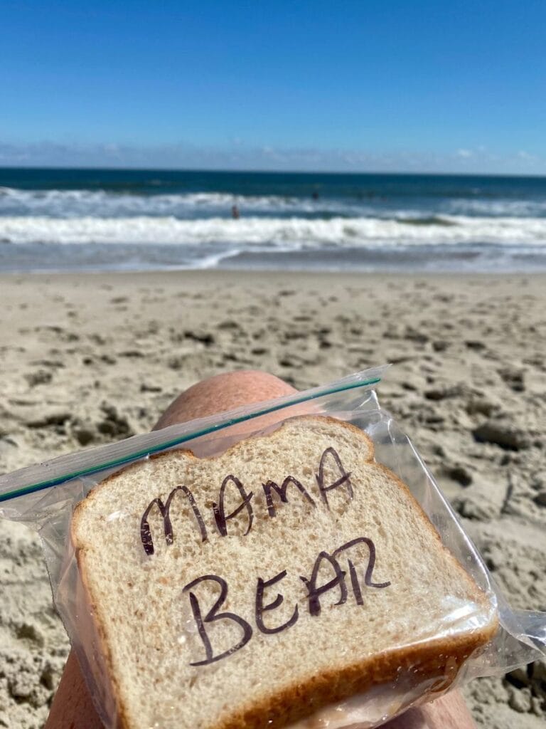A close-up of a sandwich labeled "Mama Bear" in a clear bag, with waves crashing along Kure Beach, NC, in the background.