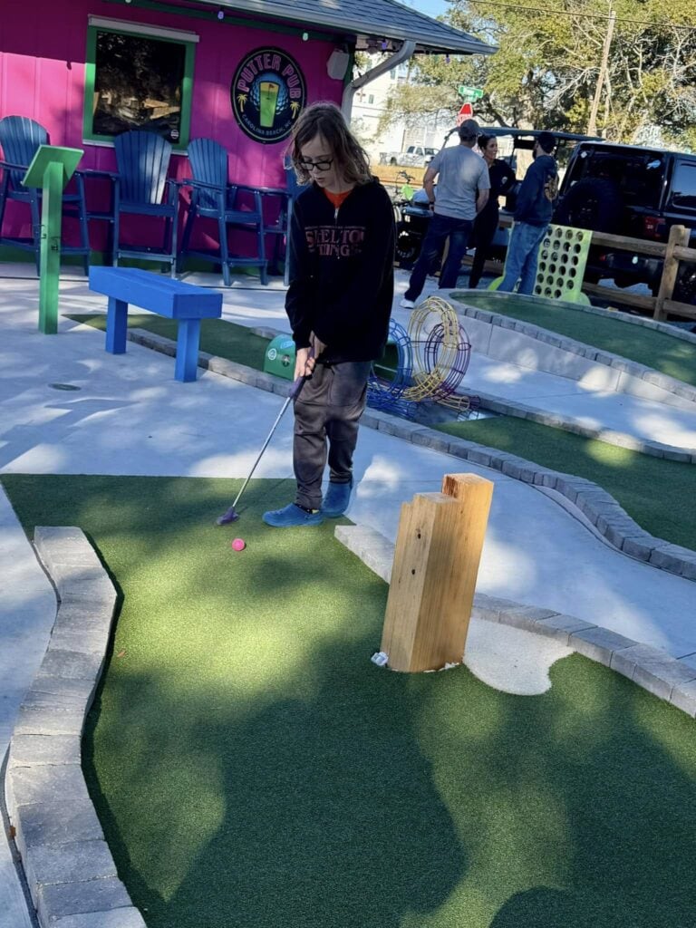 A child concentrating while playing mini-golf at the Putter Pub near Kure Beach, NC, surrounded by colorful and whimsical obstacles."