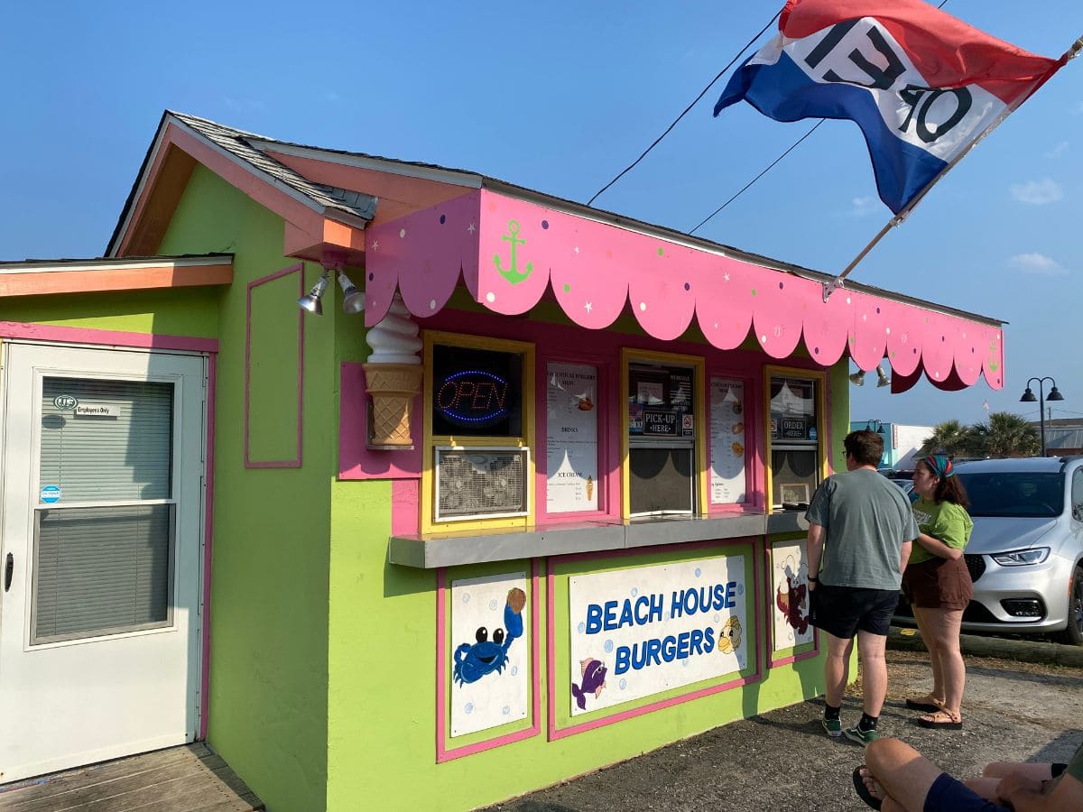 Brightly painted green and pink Beach House Burgers stand in Kure Beach, NC, with customers placing their orders under a whimsical awning featuring nautical decor and a colorful open sign.