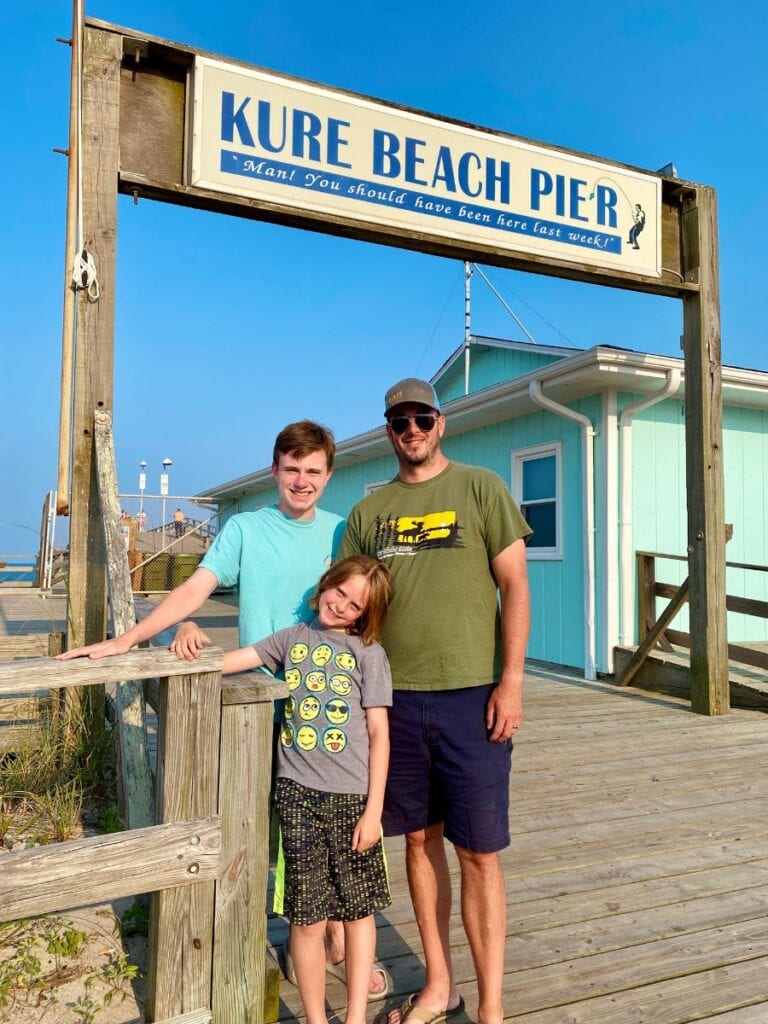 A smiling family of three posing under the sign for Kure Beach Pier, which humorously reads, "Man! You should have been here last week!"