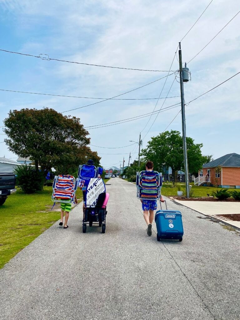A group carrying beach chairs, a cooler, and supplies down a quiet street in Kure Beach, NC, preparing for a fun day by the ocean.