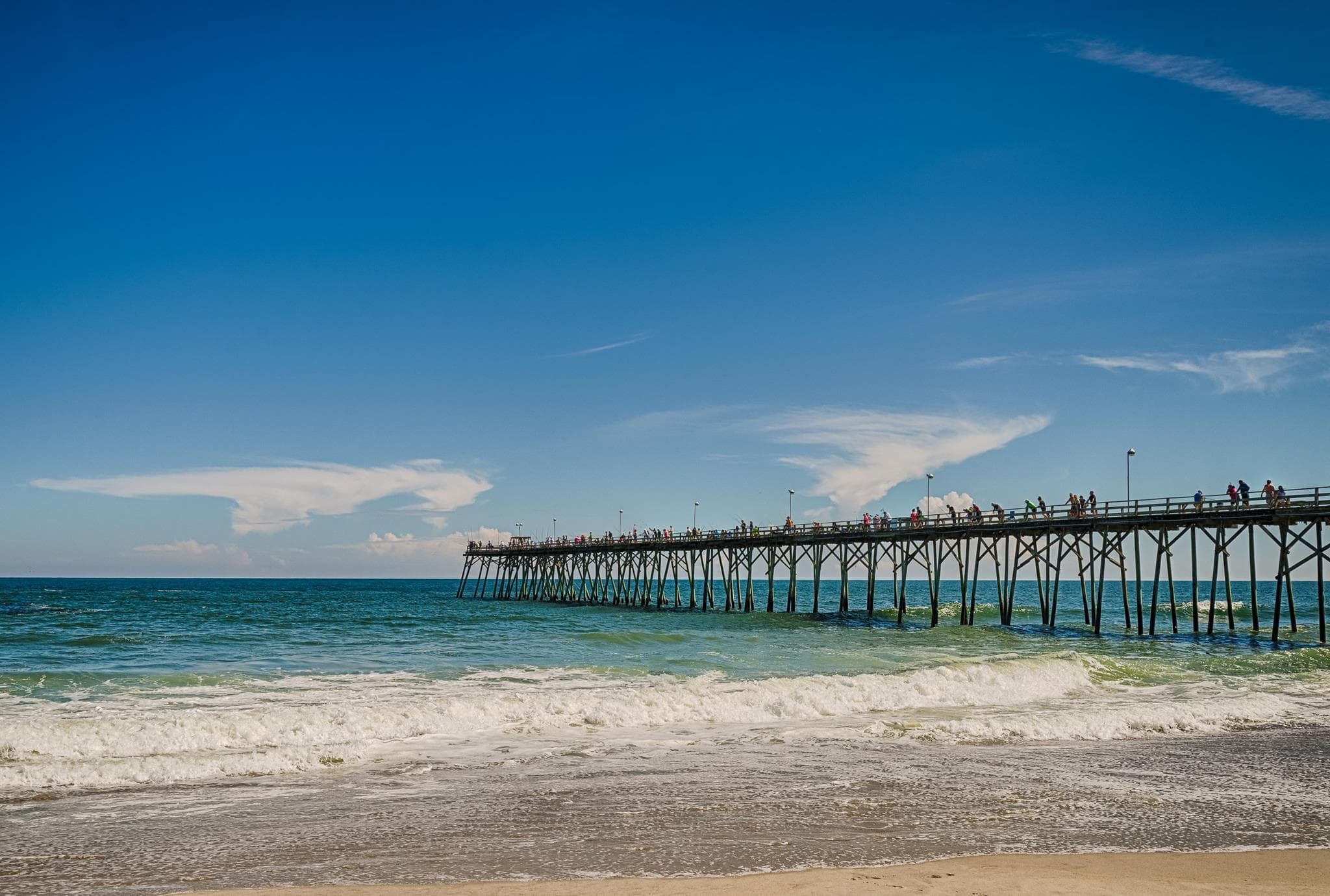 A long wooden pier stretches over turquoise ocean waves at Kure Beach, North Carolina, with people walking and fishing along its length. Bright blue skies and scattered clouds frame the scenic view, showcasing one of the top things to do in Kure Beach.